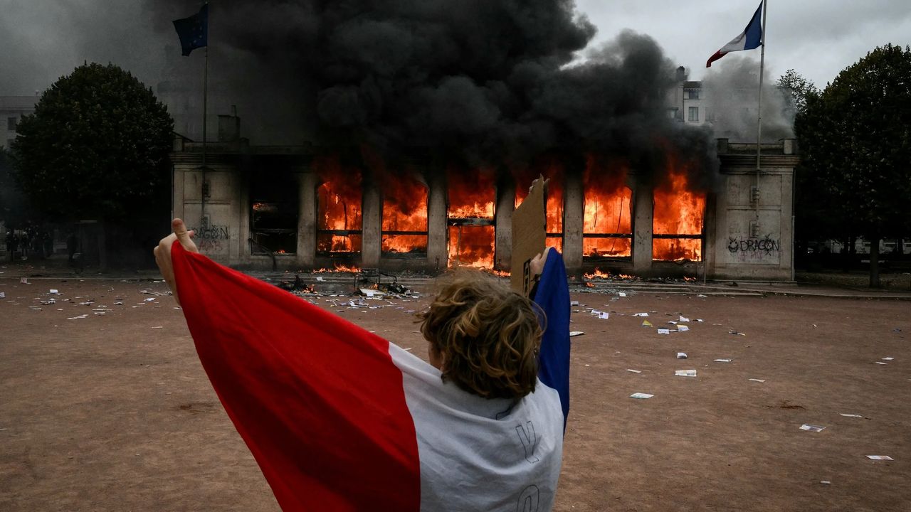 Una persona envuelta en una bandera francesa frente a un edificio público en llamas